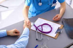 A doctor and patient shaking hands over a desk with a clipboard, form, and stethoscope